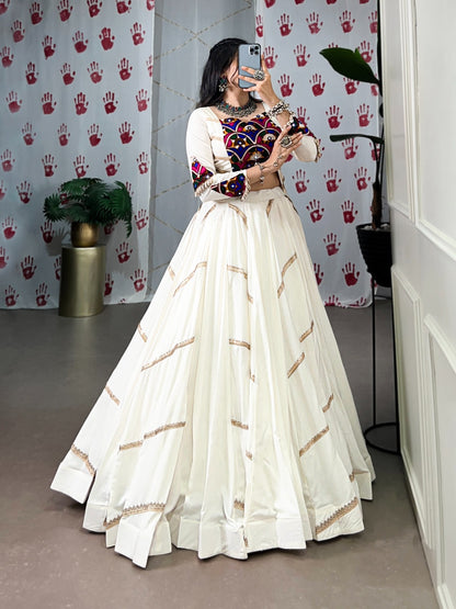 Woman in a white and gold traditional outfit with colorful top, standing in a room with patterned wallpaper.